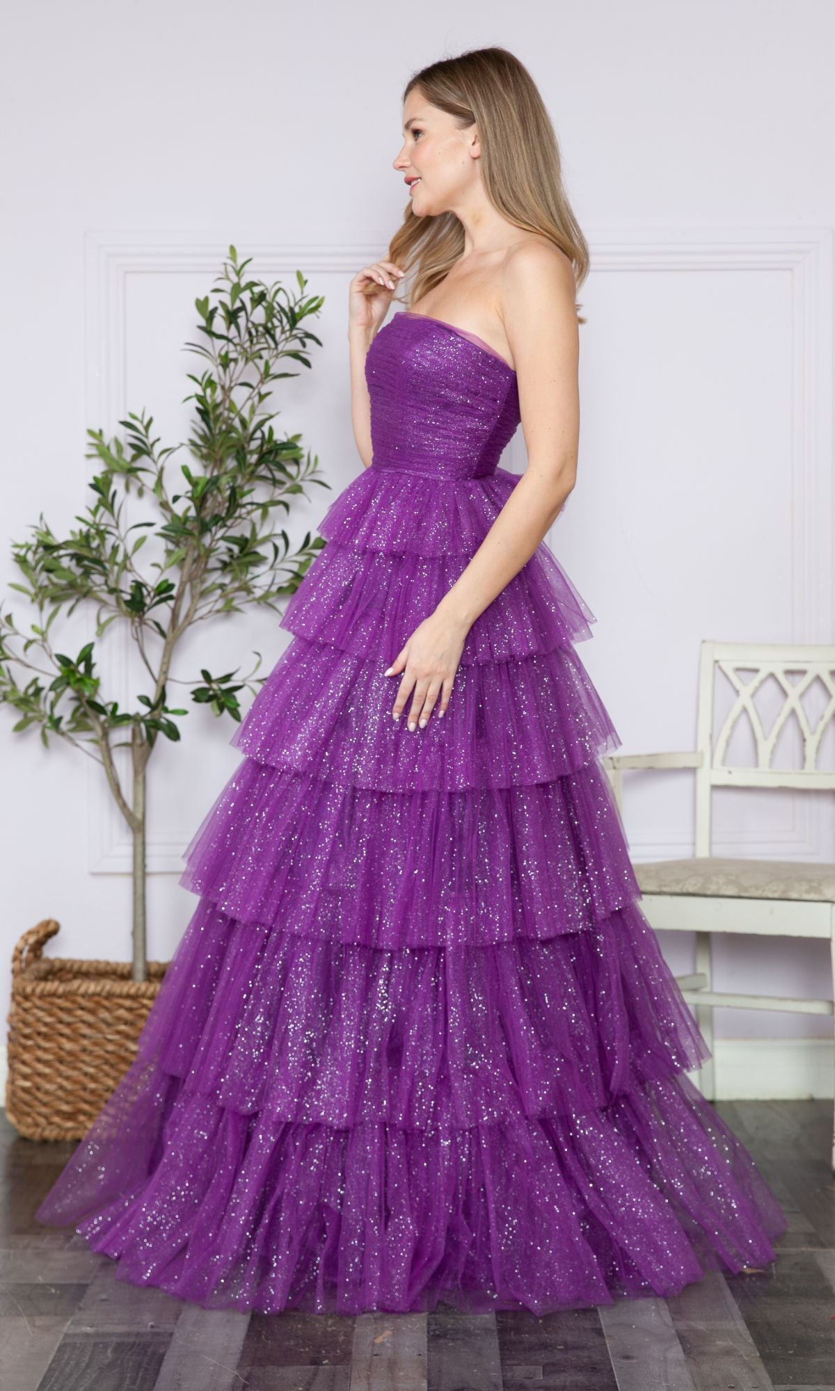 A woman stands indoors wearing the Strapless Long Tiered Glitter Prom Ball Gown 9386—a floor-length purple tulle dress. A potted plant and a white chair are in the background.